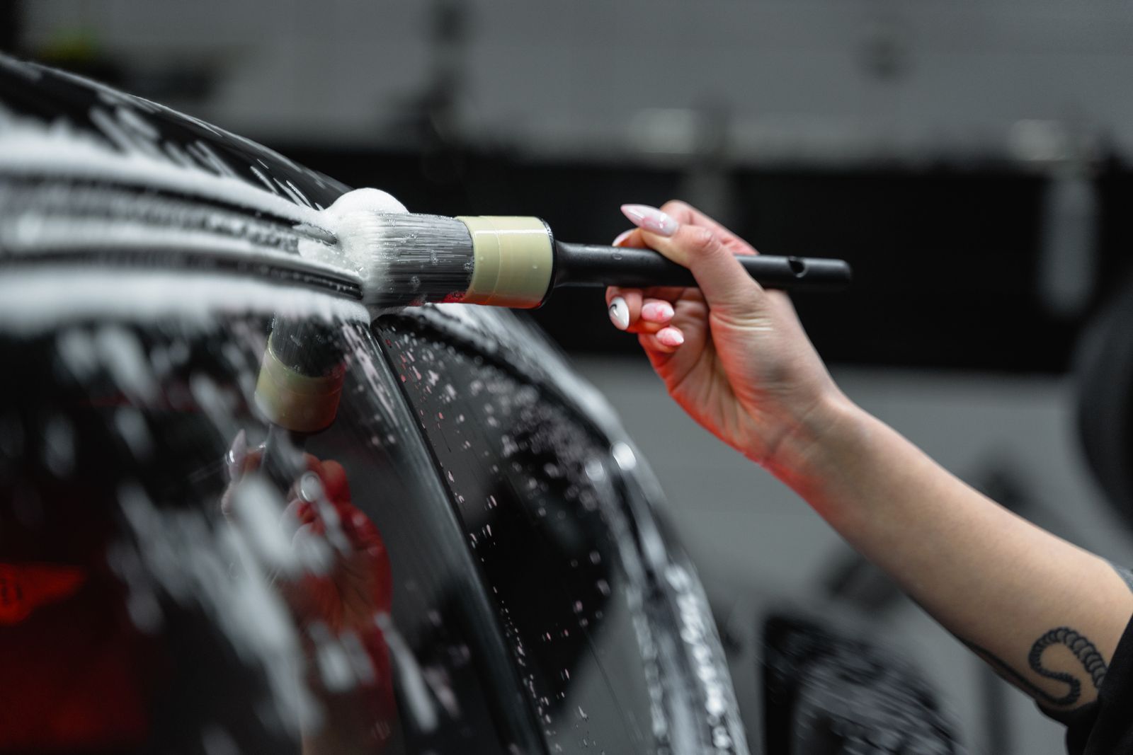 Close-up of a hand using a detailing brush to work foam into a glossy black car's exterior panel and window trim